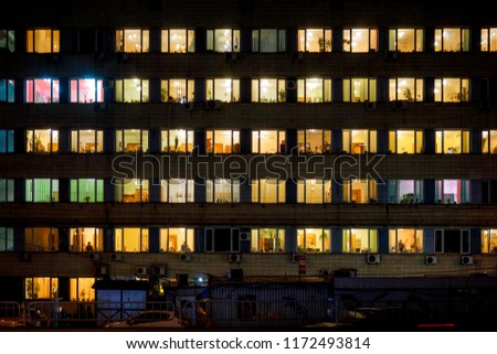 Frontal view of the night facade of building with a lot of windows with yellow green white light. life in every window. Royalty-Free Stock Photo #1172493814