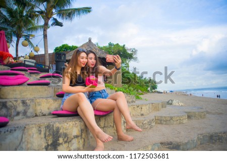 lifestyle portrait of two young happy and attractive Asian Chinese and Korean women hanging out , girlfriends enjoying holidays trip in tropical resort taking selfie picture with mobile phone 