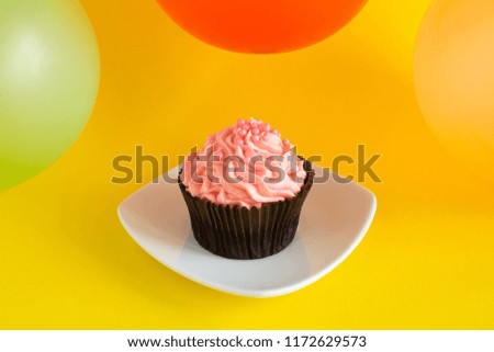 One cupcake with pink cream and colorful balloons on the yellow background