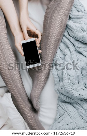 Flatlay of woman's legs in grey stocking holding smartphone with black screen in bed