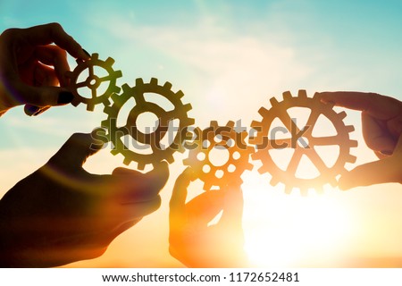 Four hands of businessmen collect gear from the gears of the details of puzzles. against the sunset. The concept of a business idea. Teamwork, strategy, cooperation, innovation. Royalty-Free Stock Photo #1172652481