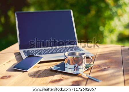 Metal steel cup of coffee and a laptop notebook on wooden table with green garden background. Freelance work place outdoors ready for coffee time.