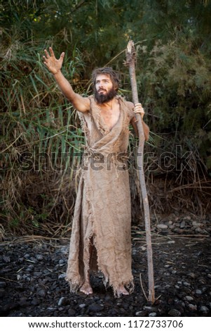 White Male Dressed as Jesus in Jewish Attire. Others as John the Baptist/ Prophet with burlap clothes and wood staff near river. Royalty-Free Stock Photo #1172733706