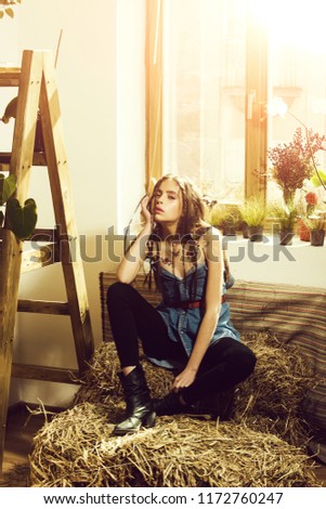 woman hippie, fashionable model, with long hair and braids in jean dress and boots sitting on natural hay at window on rural background. Country style and denim fashion. Freedom