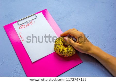 Satiric picture about diet lifestyle. Fuchsia clipboard with a “diet list” and hand holding fat yellow donut at vintage blue background. Copy space place