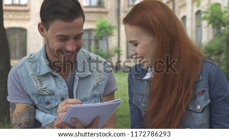 Attractive brunette male student writing something in his notebook. Close up of pretty redhead girl and handsome tatooed guy pleasantly chatting on the bench. Cute caucasian young woman smiling for