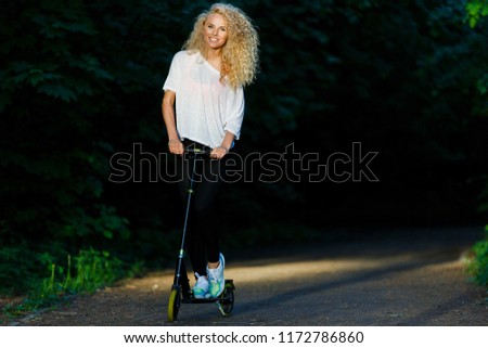 Full-length image of curly-haired athletic woman kicking on scooter in park