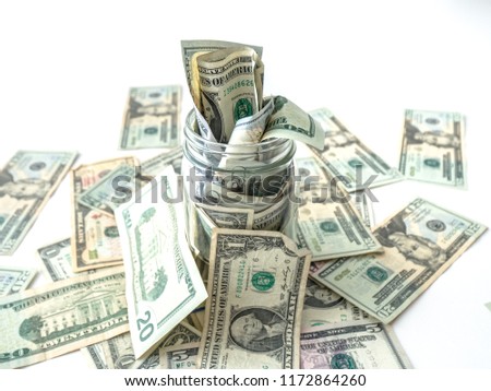 A closeup photograph of United States cash or paper bills overflowing out of a glass mason savings jar and onto a white background.
