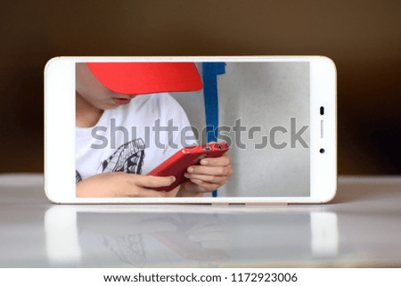 On the screen of a white smartphone is a teenager in a red baseball cap and with a red smartphone in his hands. Concept - game on a smartphone, virtual world, game addiction