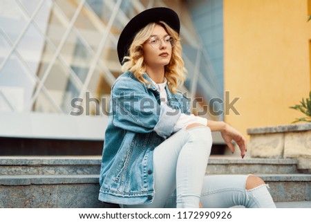 Close-up portrait of interesting woman in glasses with hat on urban background. Photo of fashionable girl with beautiful blonde hair posing while sitting on stairs.