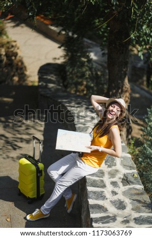 Happy laughing traveler tourist woman in yellow clothes hat with suitcase city map sitting on stone in city park outdoor. Girl traveling abroad to travel on weekend getaway. Tourism journey lifestyle