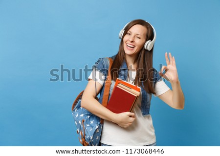 Young cheerful woman student in denim clothes with backpack headphones listen music hold school books showing OK sign blinking isolated on blue background. Education in high school university college