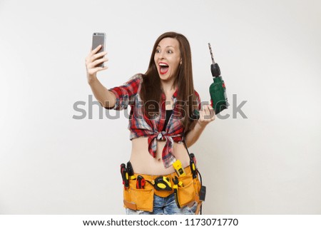 Strong excited handyman woman in shirt, kit tools belt full of instruments doing selfie on mobile phone with power electric drill isolated on white background. Female in male work. Renovation concept