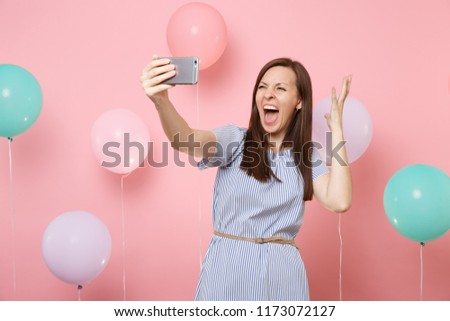 Portrait of crazy young woman in blue dress doing selfie on mobile phone screaming spreading hands on pastel pink background with colorful air balloons. Birthday holiday party people sincere emotions