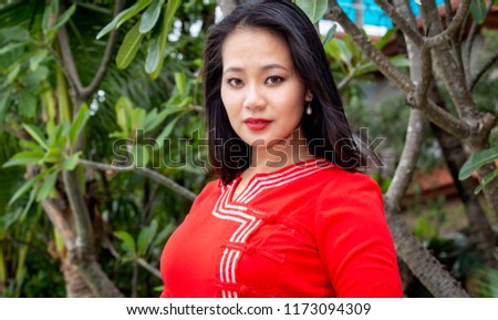 Portrait of a young Asian woman wearing a traditional Burmese red dress. Behind her some plants