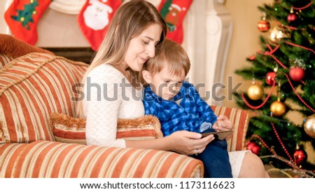 Beautiful mother sitting in armchair with her little boy next to Christmas tree and watching video on smartphone