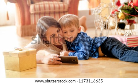 Portrait of beautiful mother lying on floor with her toddler son and watching cartoons on smartphone