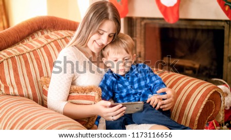 Beautiful smiling young woman resting in armchair with her little boy and watching cartoons