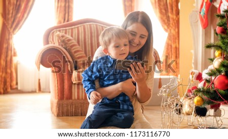 Smiling cheerful toddler boy sitting with mother under Christmas tree and watching video on smart phone