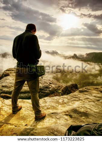 Photographer bend head check display of camera. Man stay on cliff and takes photos. Autumn beautiful misty landscape misty sunrise at horizon