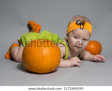 beautiful baby girl in a green dress with orange bow on her head lying on his stomach on a grey background including pumpkins