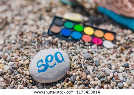 watercolor paints. Girl painting on a beach by watercolor paints. Artist traveler works on the beach. Pebble with sign sea. 