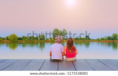 Boy with flat cap and little girl are sitting on pier. Boy and girl are looking on golden sunset. Love, friendship and childhood concept. Beautiful romantic sunset picture. 