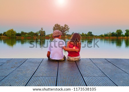 Boy with flat cap and little girl are sitting on pier. Boy is giving a hug his little sister. Love, friendship and childhood concept. Beautiful romantic sunset picture. Copy space. 