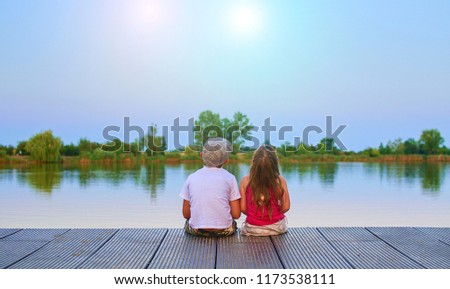 Boy with flat cap and little girl are sitting on pier. Boy and girl are looking upwards on sky. Love, friendship and childhood concept. Beautiful romantic sunset picture. 