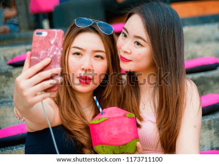 lifestyle outdoors portrait of young happy and beautiful Asian Chinese girls taking selfie picture with mobile phone enjoying holidays trip together sitting at  resort drinking tropical juice