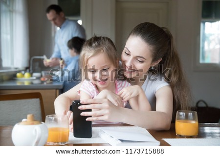 Happy mother and daughter having fun playing game on smartphone in kitchen, mom and girl spending time together watching cartoons or children video on phone, parent teach little kid using cell