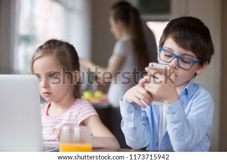 Boy and girl busy using gadgets in kitchen, siblings watch video or cartoon on laptop and play games on smartphone, small kids addicted to new technology, children having fun with devices