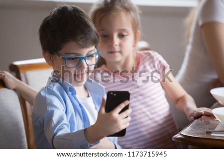 Small boy holding smartphone making picture or playing game, curious little girl looking at video on phone shown by brother, cute kids having fun watching cartoon on mobile during breakfast in kitchen
