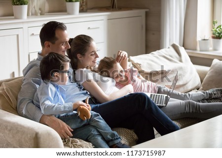 Happy parents hug kids lying on cozy sofa watching cartoons on laptop, young family spend time with children relaxing at home, mom and dad cuddle with son and daughter enjoying funny video at computer