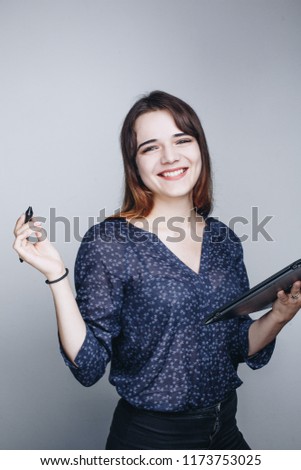 Portrait of young beautiful woman in blue blouse with tablet pen and graphic tablet in hands smiling while standing against grey background.