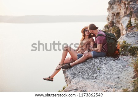 Hugging couple with backpack sitting on top of rock mountain enjoying view coast a river or lake. Traveling mountains and coast, freedom and active lifestyle concept.