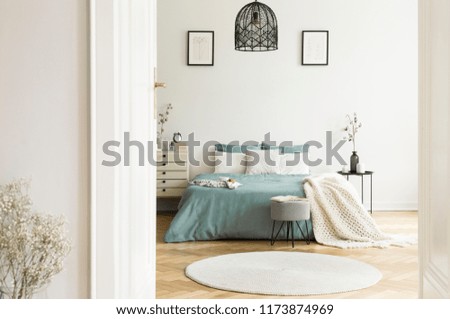 A view through an open door into a sunny bedroom interior with sage color linen and cushions on a bed, blanket, drawer cabinet, soft stool and a round rug. Real photo