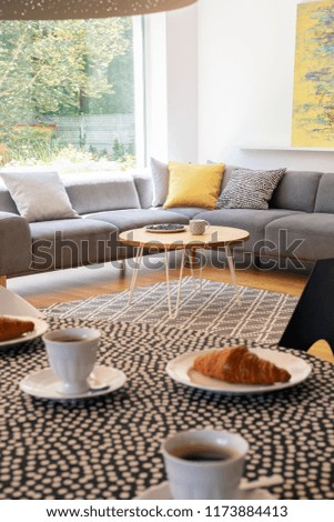 Focus on a gray corner sofa by a big window and a round table and blurry foreground with pastries and coffee in a modern coffee shop interior