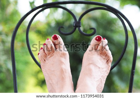 Woman's feet relaxing against wrought-iron garden chair. Brilliant red nail varnish. Close-up, selective focus, green blurred background.         