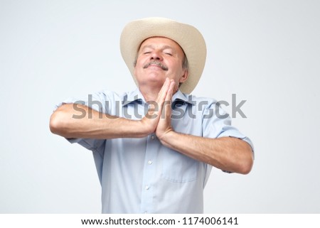 Pleased hopeful mature man closes eyes, keeps palms together, prays to have good luck, has dreamful look, isolated over gray background. Old cowboy asking for benefits from God