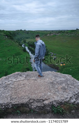 Young male photographer standing on top of the mountain, holding the camera and looking at the neighborhood, choosing a frame for shooting, the view from the back