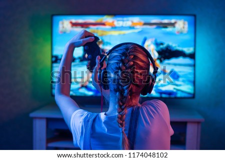 A gamer or a streamer girl at home in a dark room with a gamepad playing with friends on the networks in video games. A young man sits in front of a monitor or TV. Royalty-Free Stock Photo #1174048102