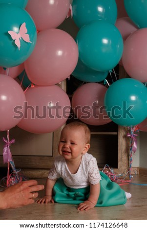 the child cries, gets angry, a little girl, a child, sits on the floor and cries, wrinkled her nose, near the balloons, a holiday. birthday, turned a year old. resentment, disappointment