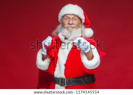 Christmas. Smiling Santa Claus in white gloves with a bag of gifts behind him points his index finger into the camera. Isolated on red background.