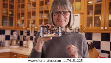 Happy white grandmother showing off video of family on mobile device in kitchen
