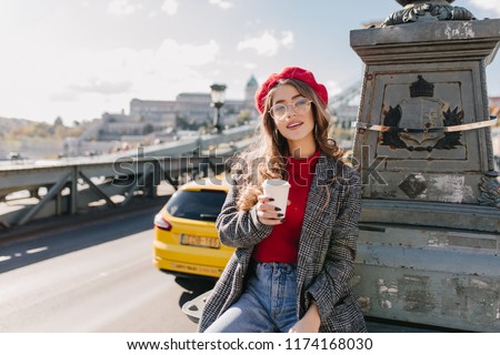 Interested brown-haired woman in blue denim pants holding cup of cappuccino on sky background in cold day. Photo of enchanting confident girl drinking coffee on the road.