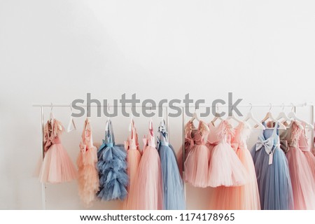 Beautiful dressy lush pink and blue dresses for girls on hangers at the background of white wall. Kids dresses with feathers for prom and holiday. Royalty-Free Stock Photo #1174178965