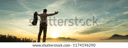 Happy successful businessman celebrating on a wall watching the sunrise silhouetted against the colorful orange sky in a panorama view with copy space. Royalty-Free Stock Photo #1174196290