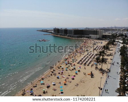 Aerial photo of the coastal beach of Alicante in Spain, the photo shows people sunbathing on the beach with parasols and people swimming in the water, you can also see hotels in the background.