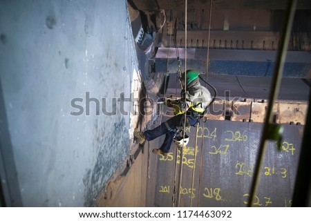 Wide angle picture of rope access sandblaster worker wearing safety equipment harness working at height abseiling on twin ropes commencing sandblasting in confined space construction site Perth, Aust 